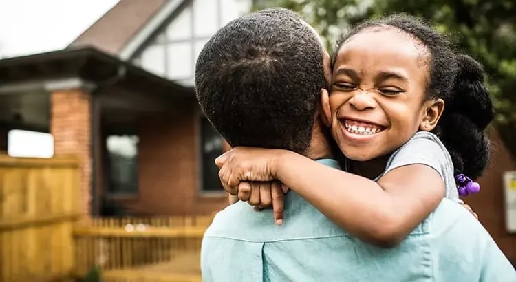 A young girl hugging an adult, likely a parent, with a joyful expression. They are outside, near a house, and the image captures a moment of affection.