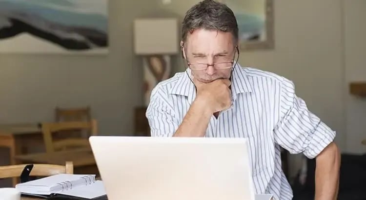 A middle-aged man is sitting at a desk, deeply focused on his laptop. The background suggests a cozy home office setting.