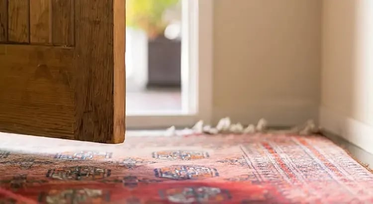 The lower part of a wooden door that is partially open, revealing a glimpse into another room. The door is made of natural wood with visible grain patterns. On the floor, there is a colorful, intricately patterned rug with predominantly red tones and various geometric and floral motifs. The edge of the rug appears slightly worn or frayed.