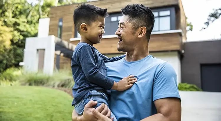 A man holding a young child in an outdoor setting, possibly a backyard or park. They are both smiling and appear to be enjoying a moment together.