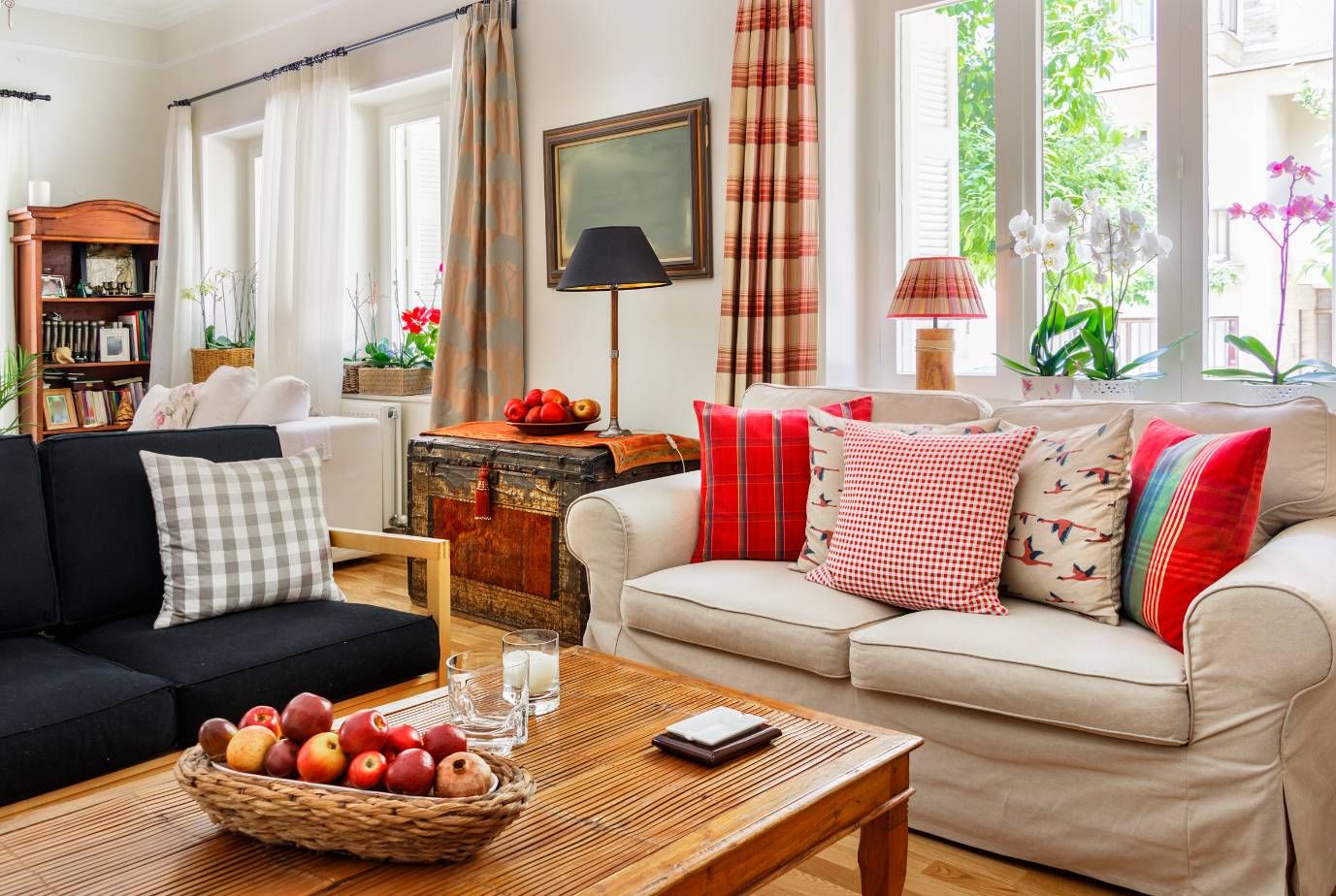 Living room with colorful plaid pillows, a basket of apples, and orchids beside tall windows.