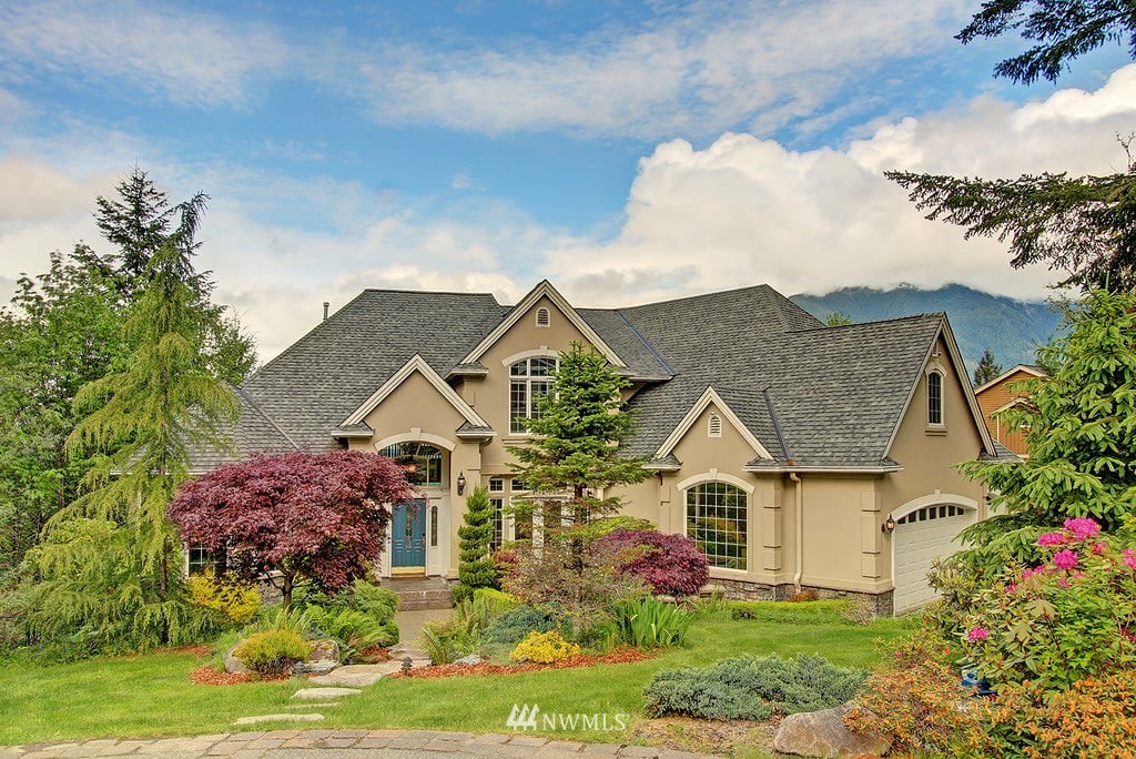 Beige home with gray gabled roofs, blue front door, landscaped garden, and mountain backdrop.