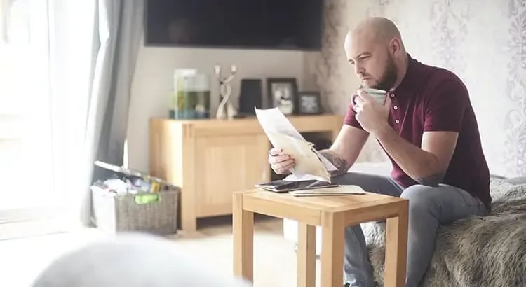 He is wearing a maroon polo shirt and light blue jeans. The man is holding a coffee cup in one hand and looking at a piece of paper in the other hand.