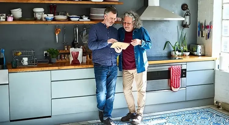Two men standing in a kitchen, discussing something. The kitchen has a modern design with gray cabinets and stainless steel appliances, indicating a warm, familial moment.