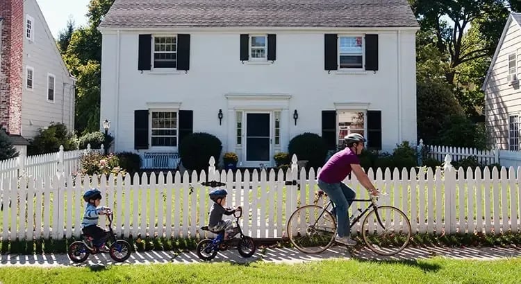 A person riding a bicycle with children on a smaller bike alongside them. They are passing by a white picket fence in front of a classic two-story house with black shutters.