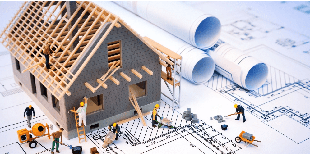House under construction on blueprints, with miniature workers assembling its roof and walls.