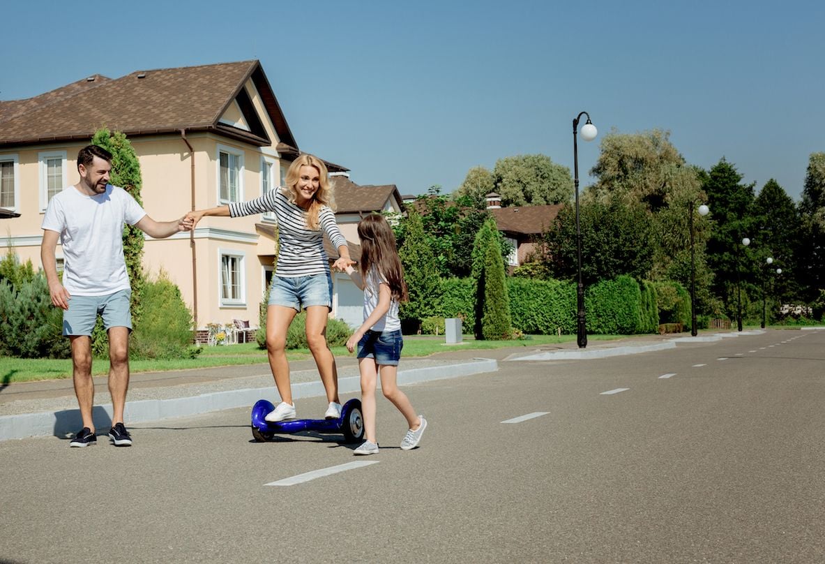 A man and girl steady a smiling woman riding a blue scooter on a residential street.