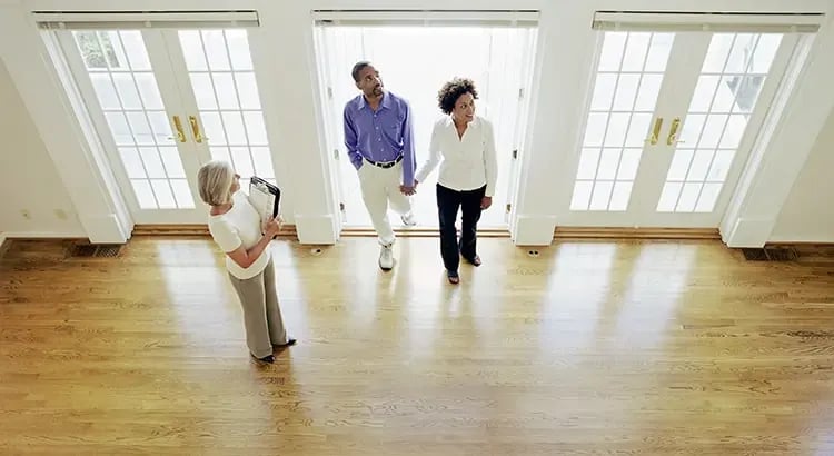 There is a couple holding hands, standing in the middle of the room.  Another woman, possibly a real estate agent, is standing to the left, holding a clipboard or tablet. 