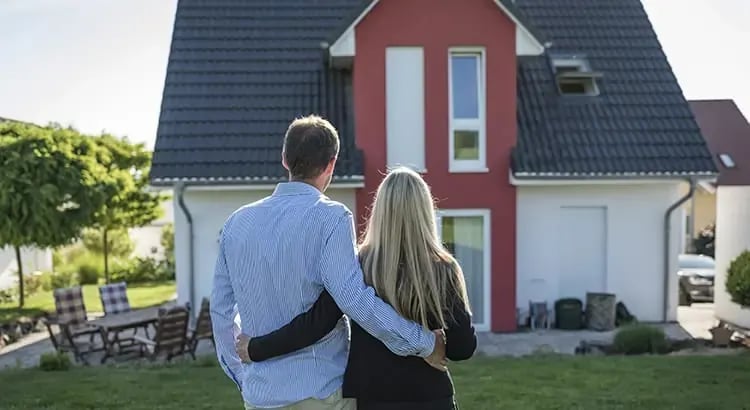 A couple standing arm in arm, looking at a house. The house is red with white trim, and the couple appears to be prospective buyers or homeowners.