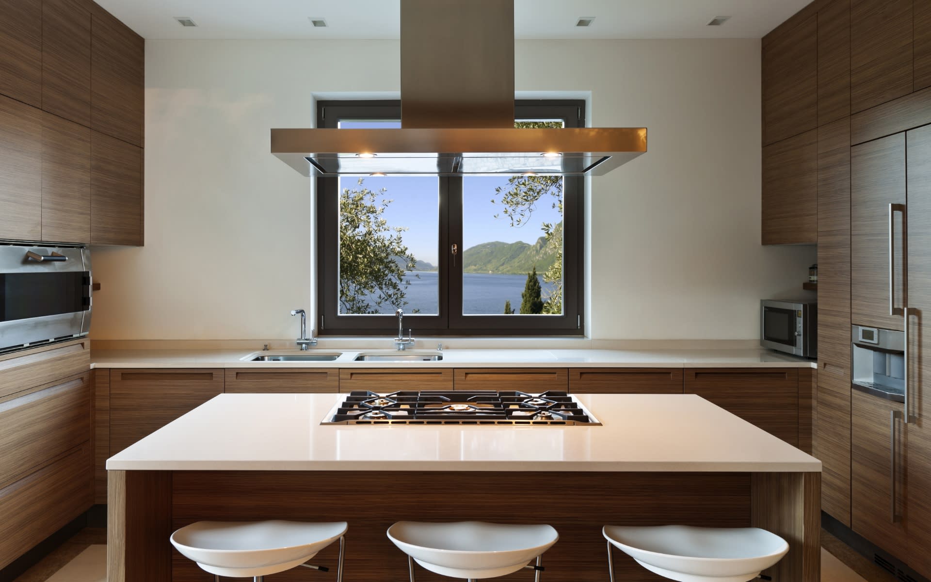 A modern kitchen with warm brown cabinets and a white island overlooks a serene blue lake with green hills in the background.