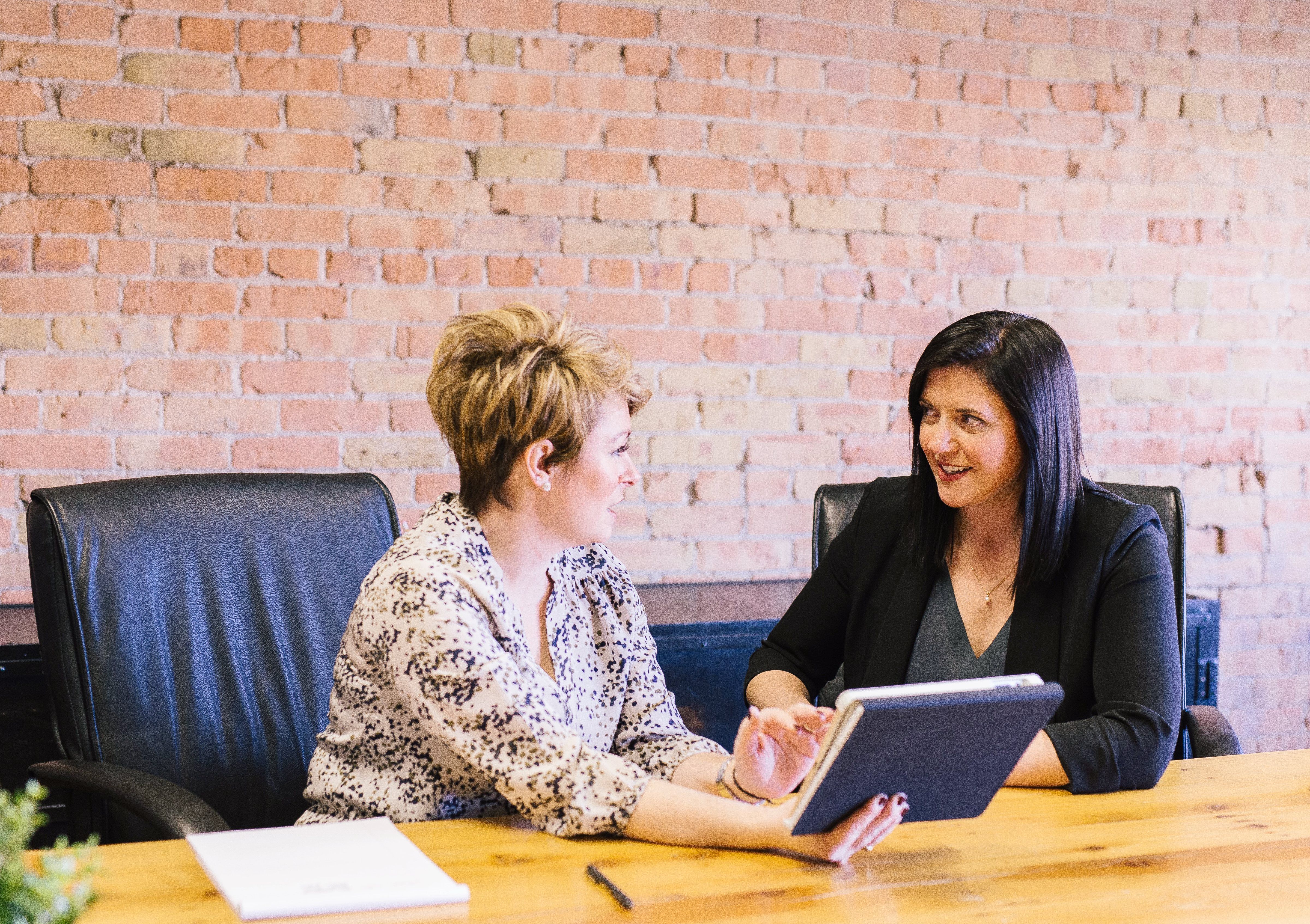 Two women discuss information on a tablet across an office table.
