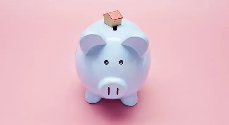 A blue piggy bank with a house-shaped object on top, set against a pink background. The image represents saving money for home ownership or financial planning.