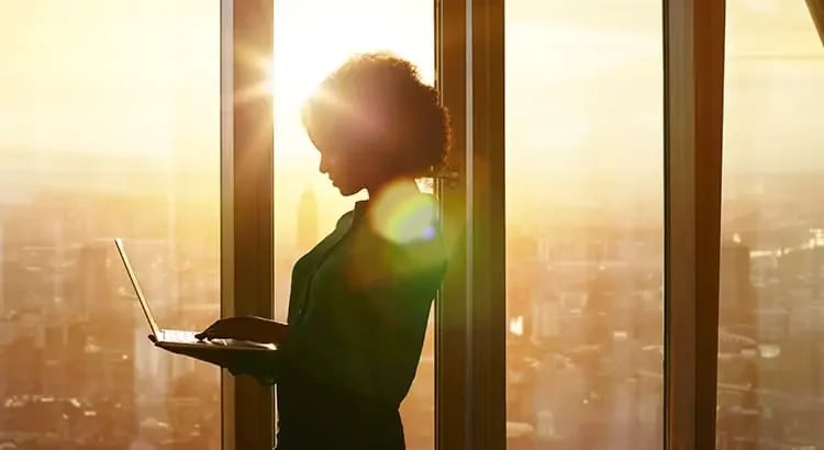 A woman standing by a large window, holding a laptop. The sunlight is streaming in, creating a silhouette effect.
