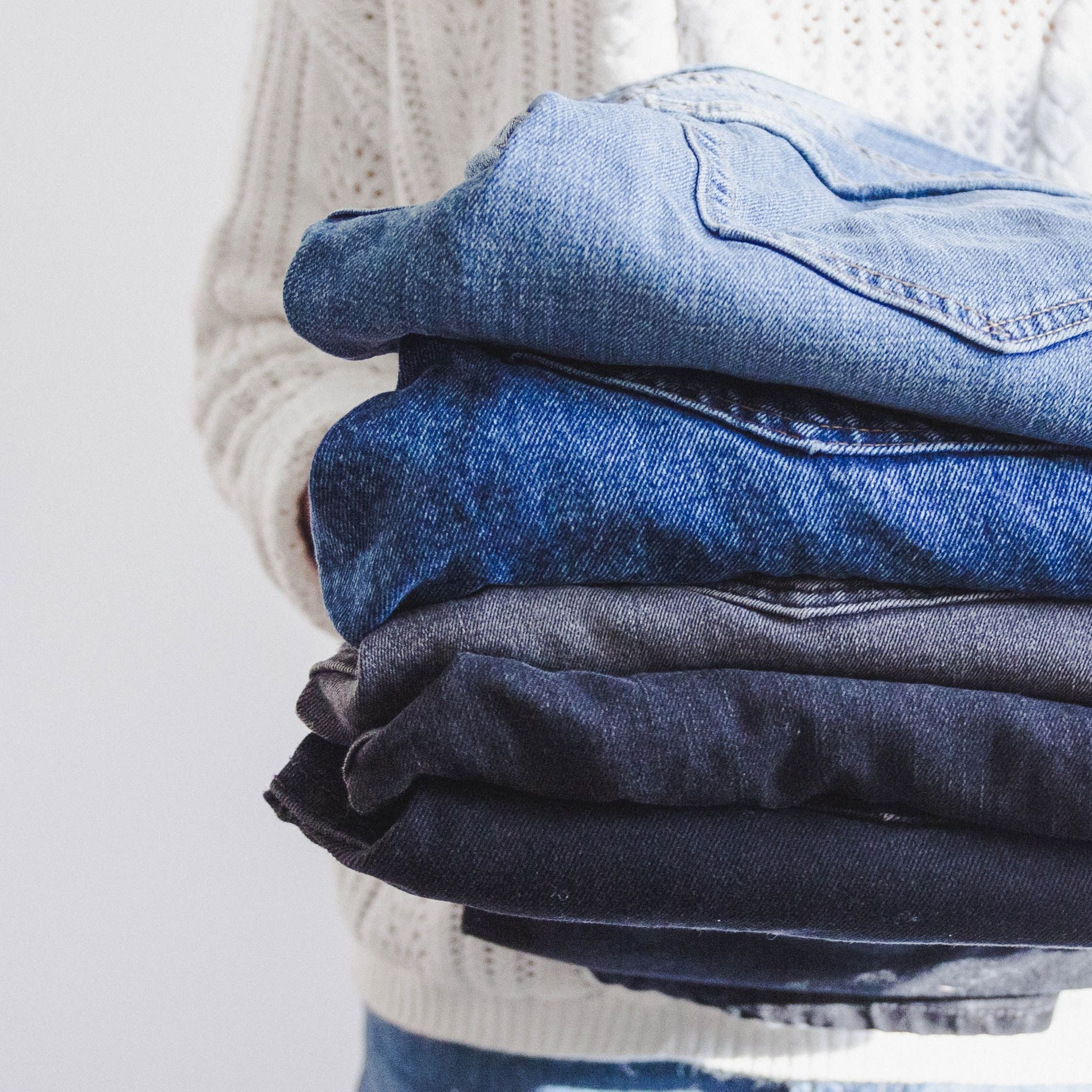 A person holds folded blue and gray jeans against a cream knit sweater.