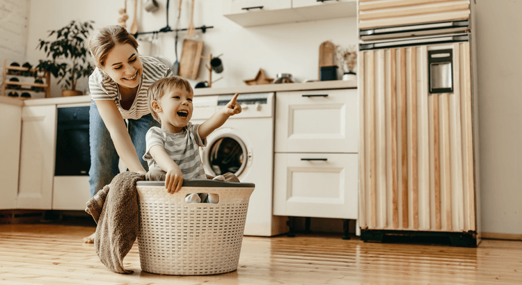 Woman and young child play in a laundry basket in a bright kitchen.