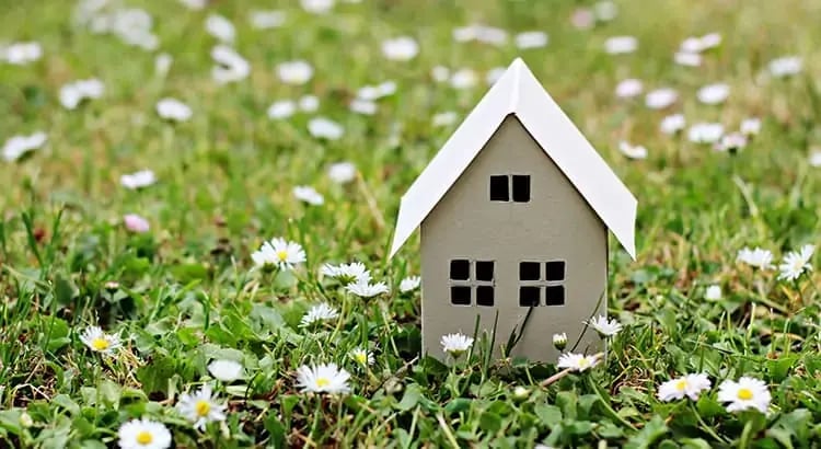 A small model house placed on a grassy field with white flowers, possibly daisies. This image symbolizes home ownership, real estate, or springtime.