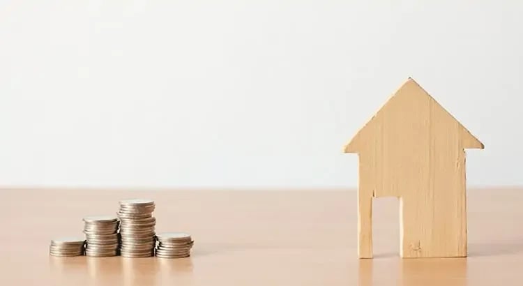 A wooden house model placed next to stacks of coins on a wooden surface, representing financial aspects of homeownership or investment.