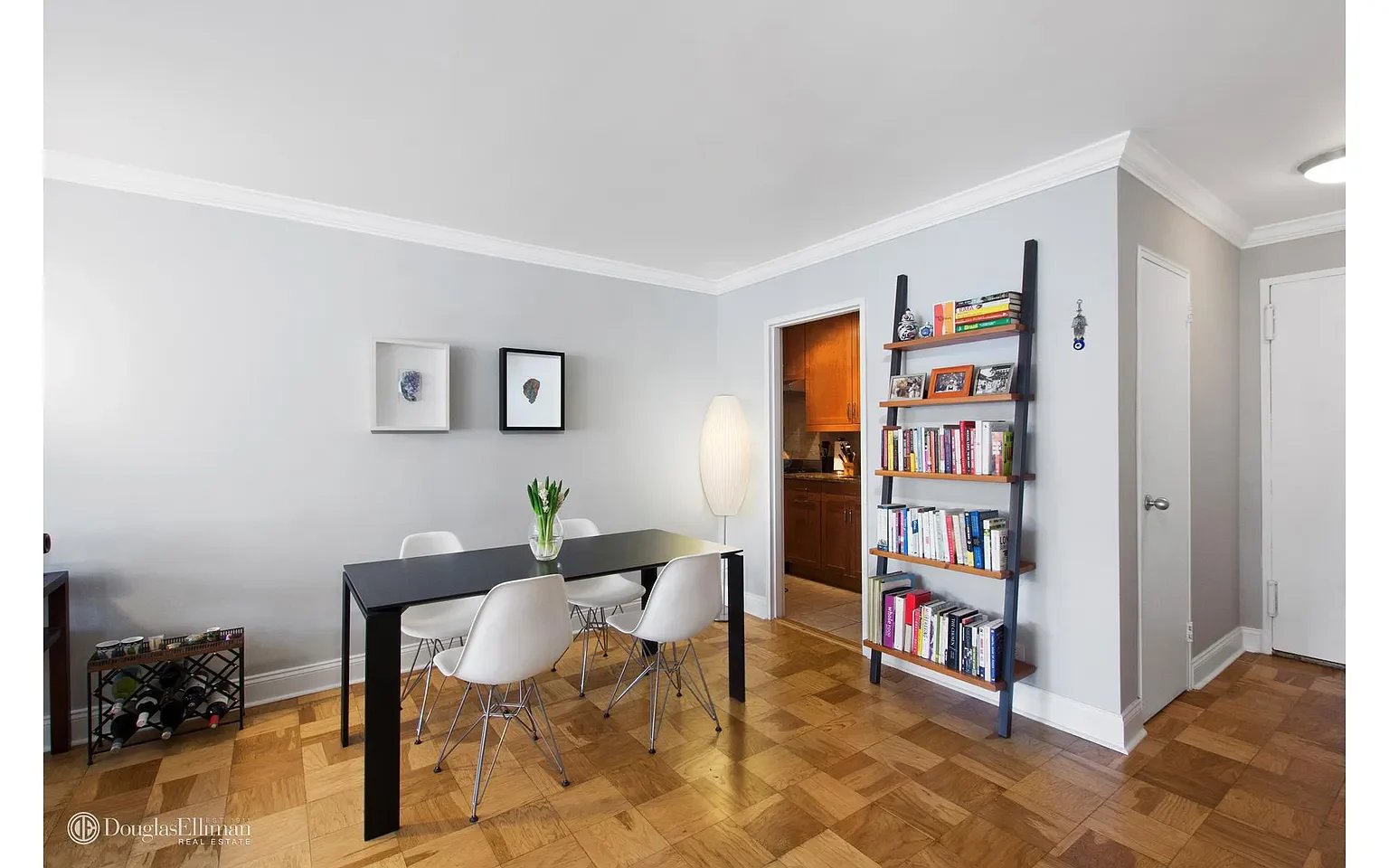 Dining area with a black table, white chairs, parquet flooring, and a tall bookcase.