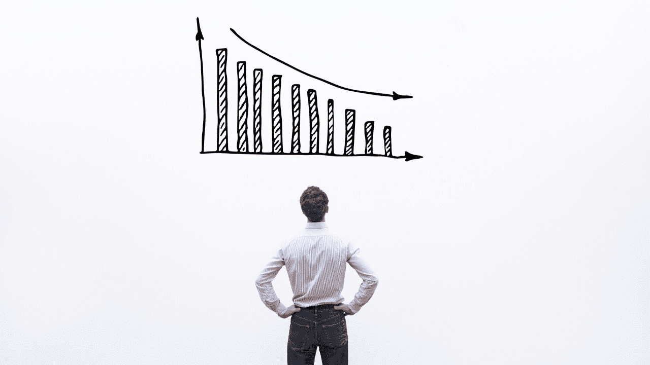 A man standing with his hands on his hips, facing a hand-drawn bar chart on a white background. The man is viewed from behind and is dressed in a formal or semi-formal outfit, wearing a striped shirt and jeans.