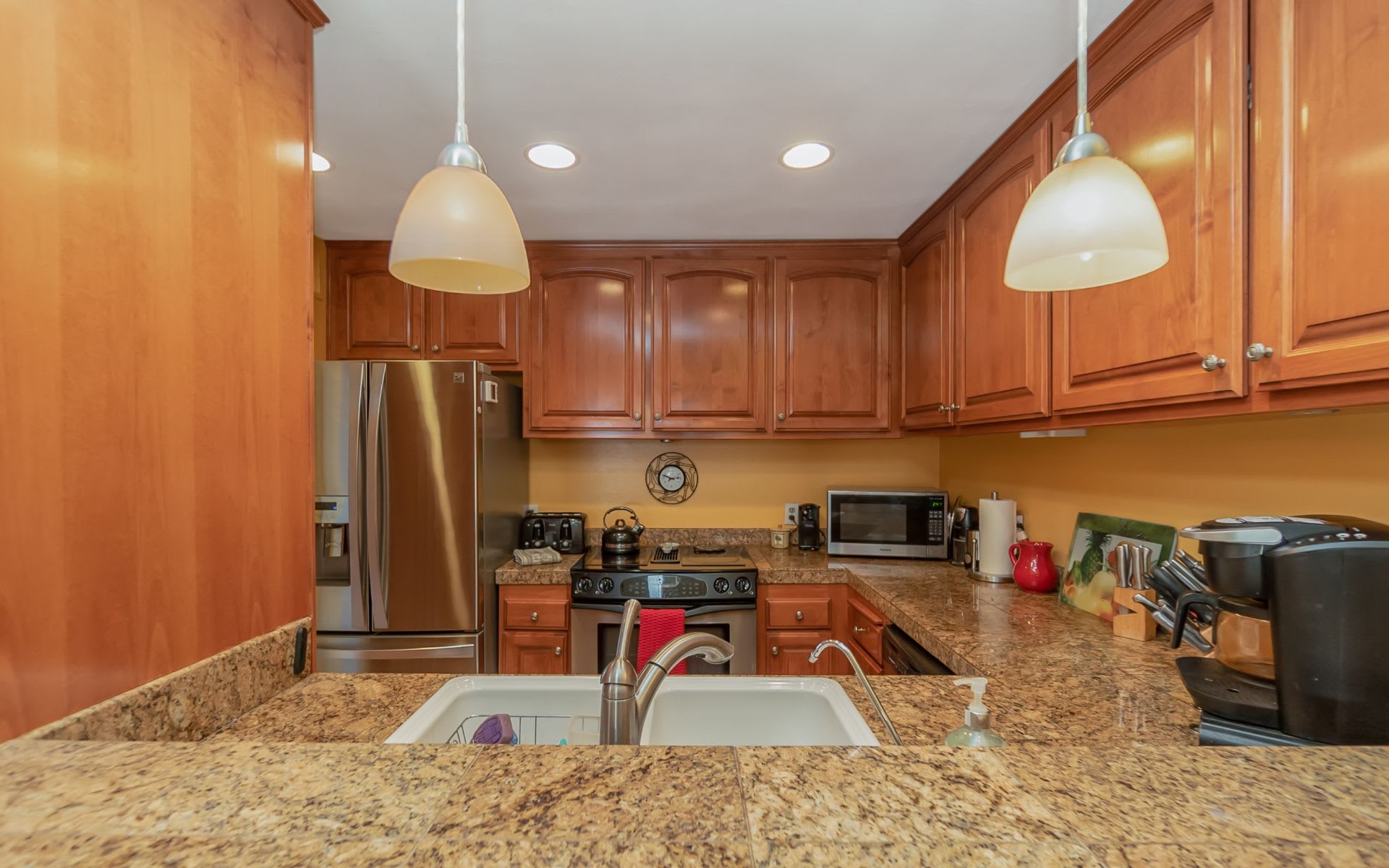 Kitchen with reddish wood cabinets, granite counters, a foreground sink, and stainless-steel refrigerator.