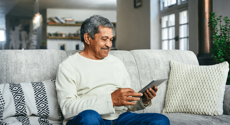 A photo of a man indoors, sitting comfortably on a couch, using a tablet. The setting appears cozy and relaxed.