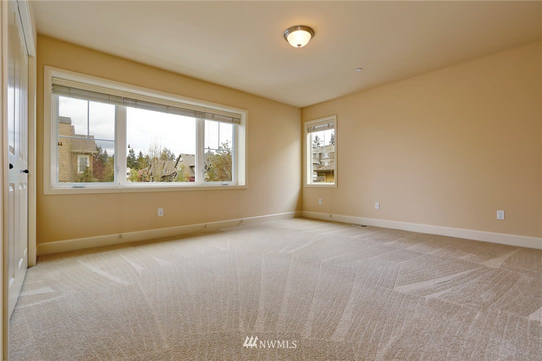 Empty bedroom with beige carpet and two windows overlooking neighboring homes.