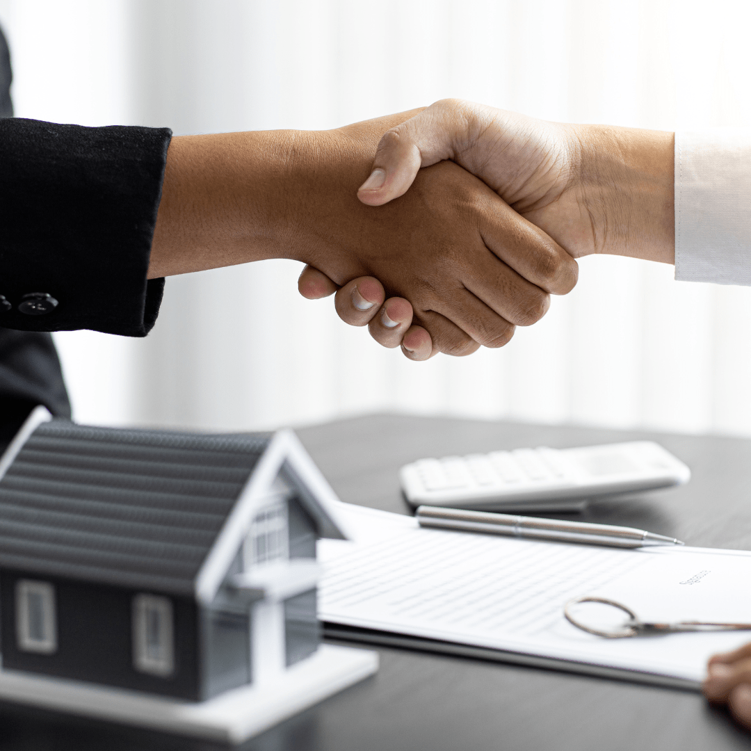 Two people shake hands over a model house and real estate documents.