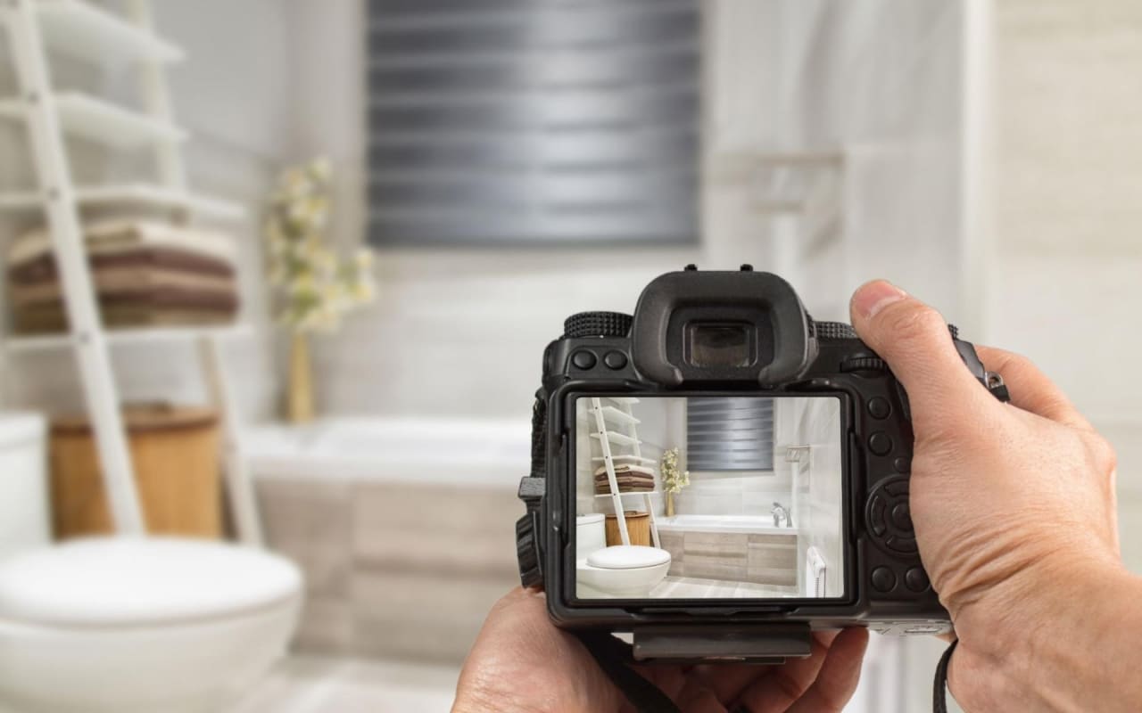 A person takes a photo of a clean, modern bathroom with white walls and a bathtub.