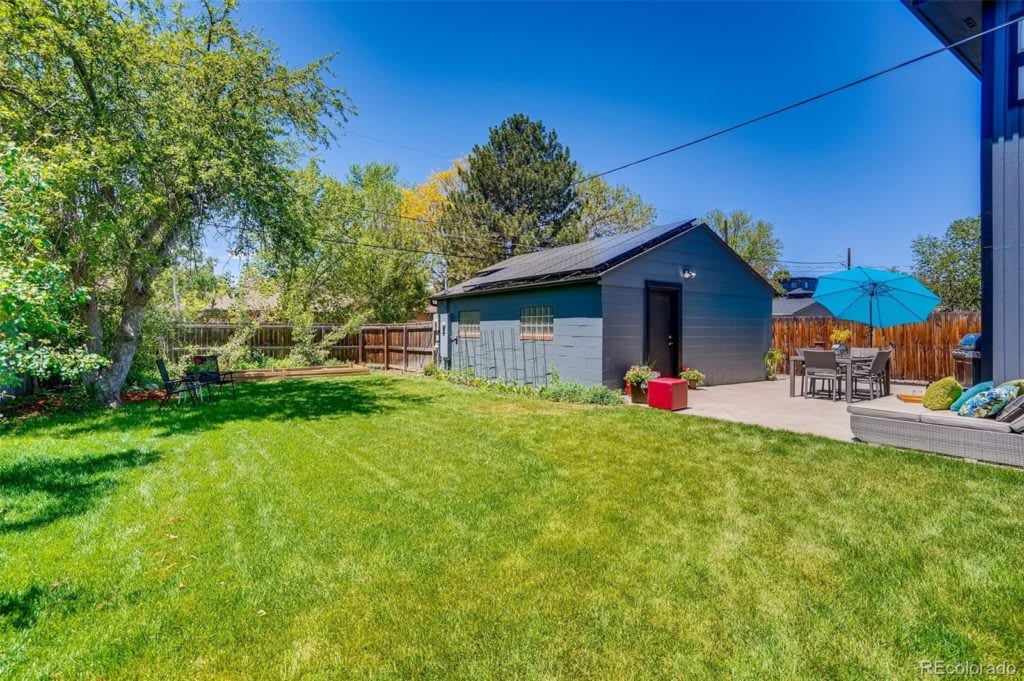 Fenced backyard with a broad lawn, detached gray outbuilding, and patio seating beneath a blue umbrella.
