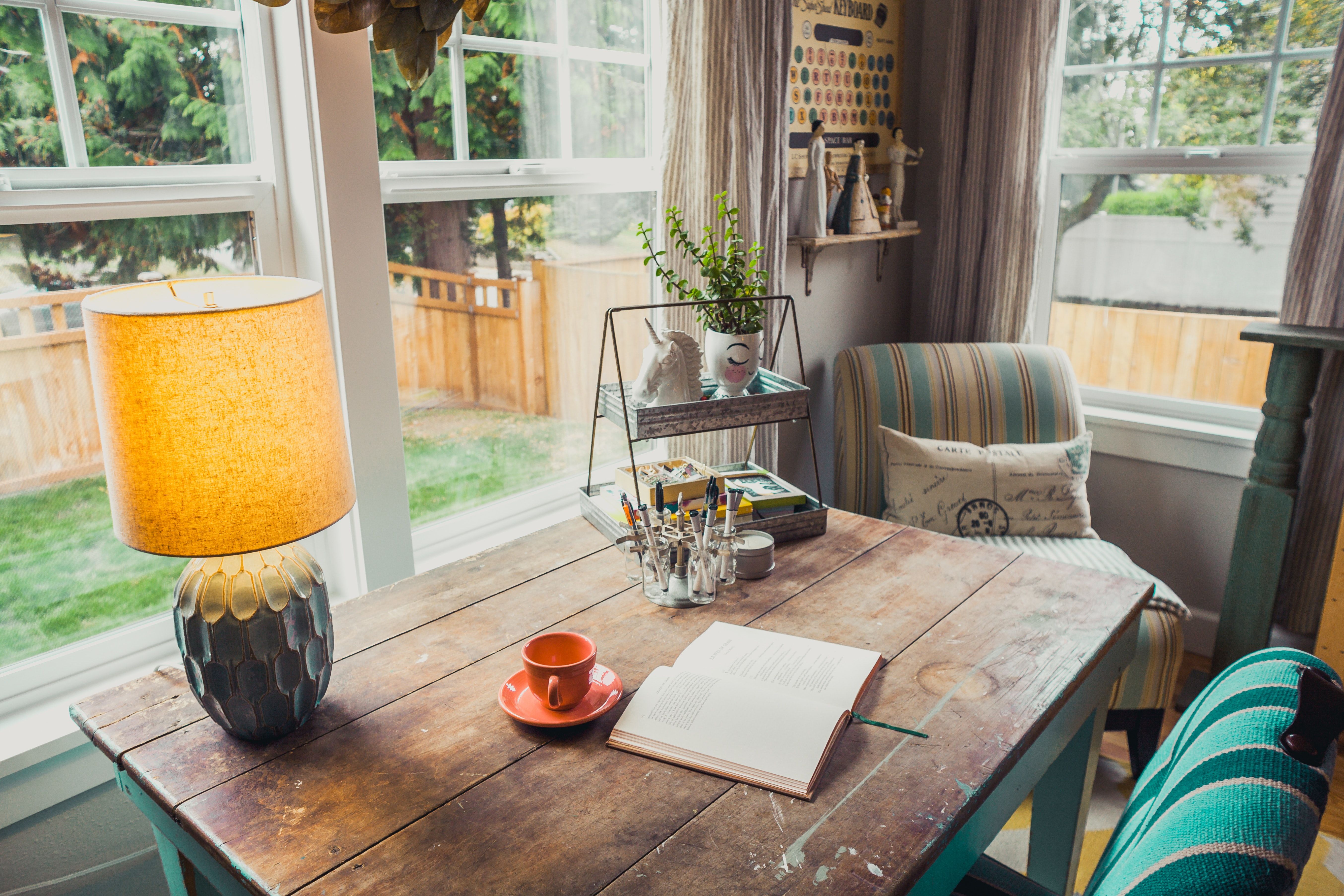 Furnished sitting area with a rustic table, open book, glowing lamp, and striped armchair beside large backyard-facing windows.