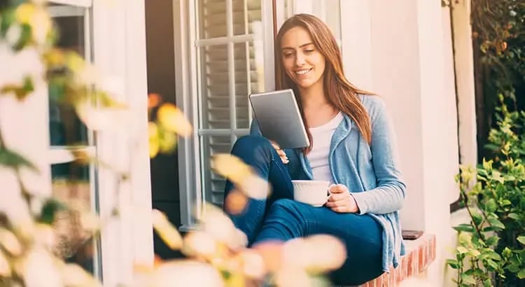 A woman sitting outdoors with a tablet, smiling and engaged with the device. The background is filled with flowers and greenery, creating a serene setting.