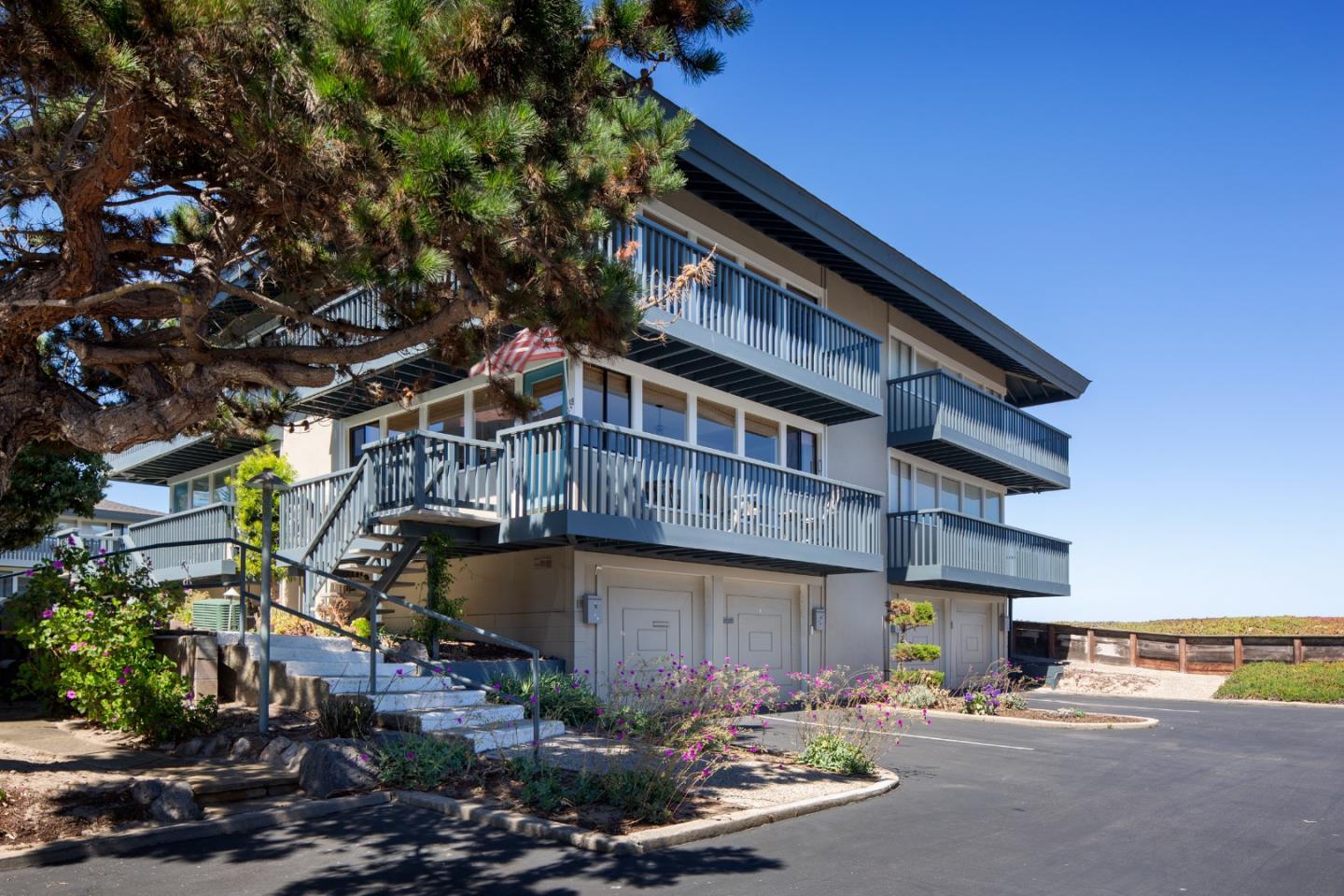 Multi-story residence with broad balconies, an exterior staircase, and a large pine tree in front.