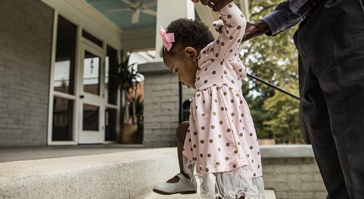 A child reaching up while standing on a step, possibly about to enter a house, assisted by an adult. The setting suggests family or home life.