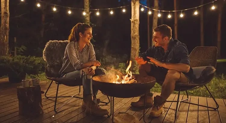 Two people sitting around a fire pit in an outdoor setting, possibly in a backyard or campsite, having a discussion.