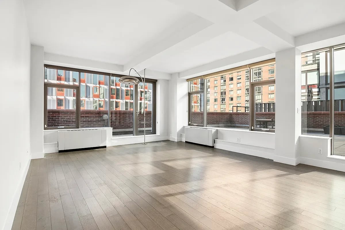 Vacant living room with pale wood floors and large windows overlooking neighboring buildings.