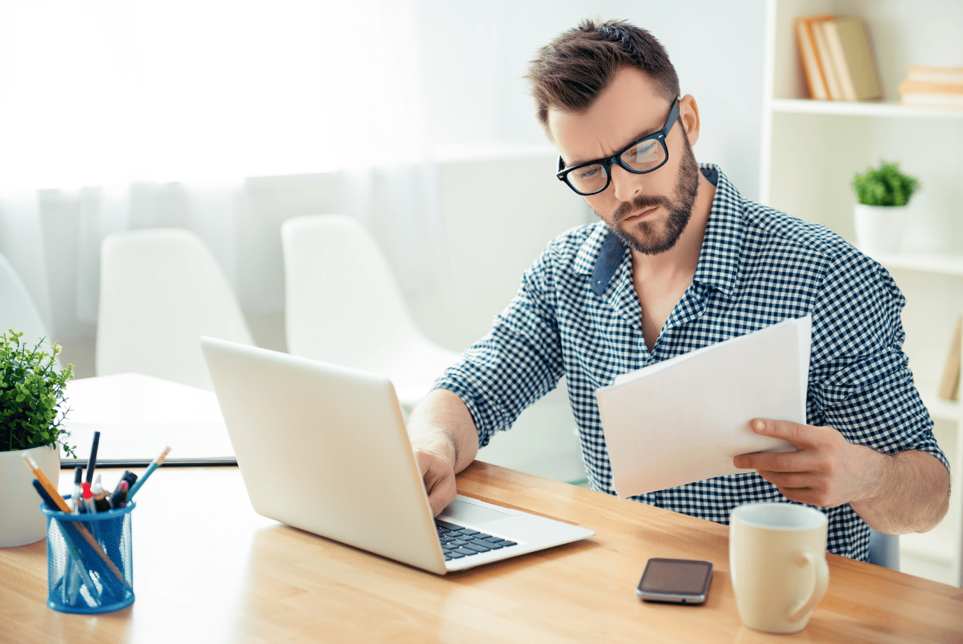 Man with glasses studies a document beside his laptop at a bright office desk.