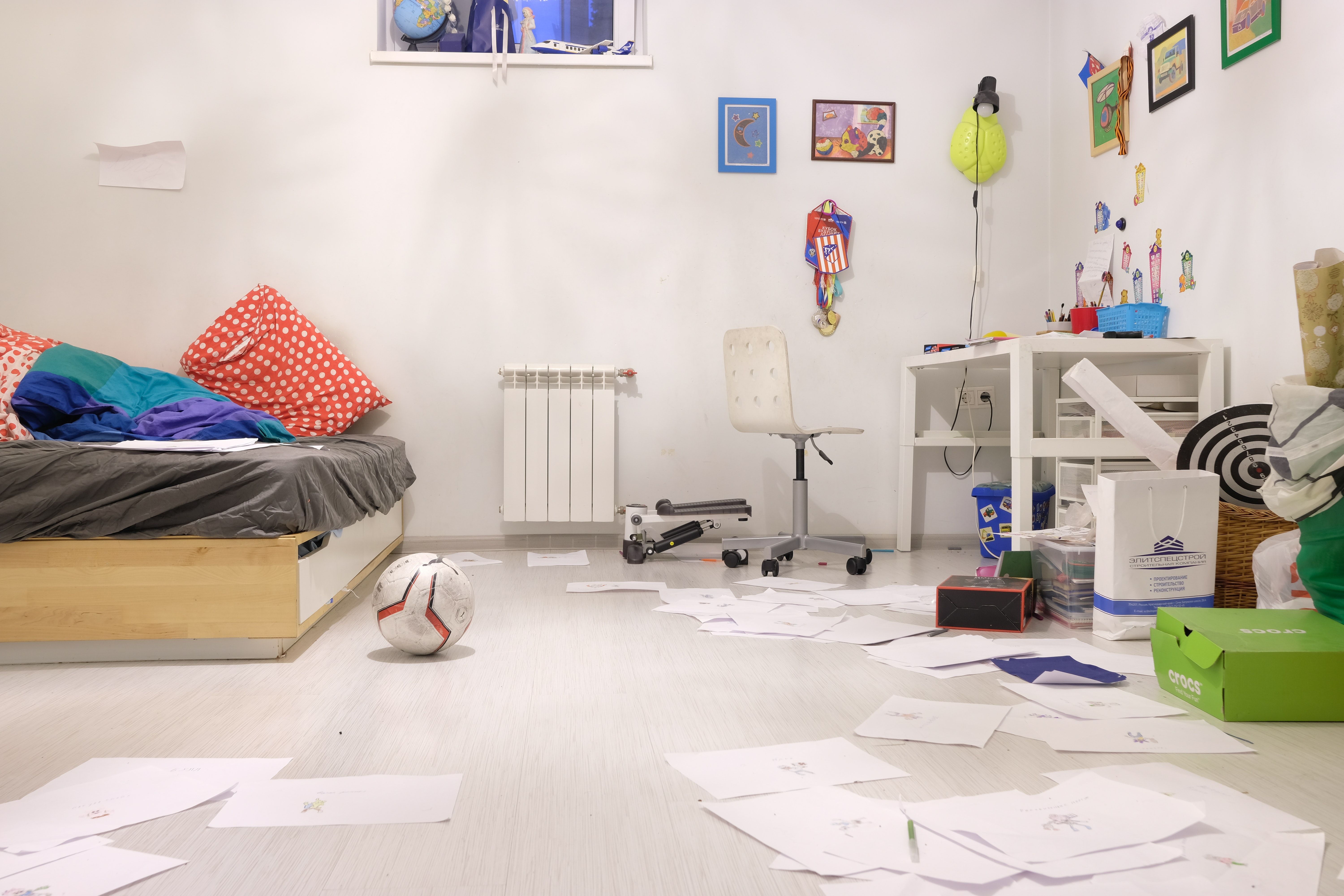 Child's bedroom with colorful bedding, a desk, sports items, and papers scattered across the floor.