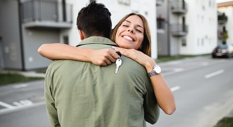 a joyful moment between two people, where a woman is hugging a man from behind, holding a key in her hand. 