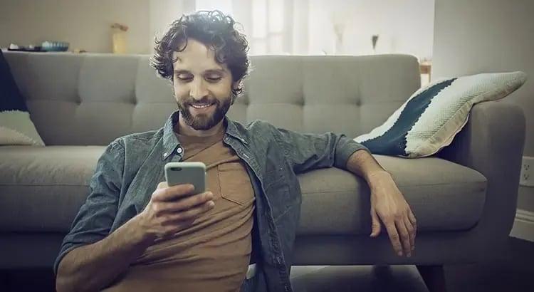 A man sitting on a couch, looking at his phone and smiling. The setting is relaxed and comfortable, indicating leisure time or social interaction.