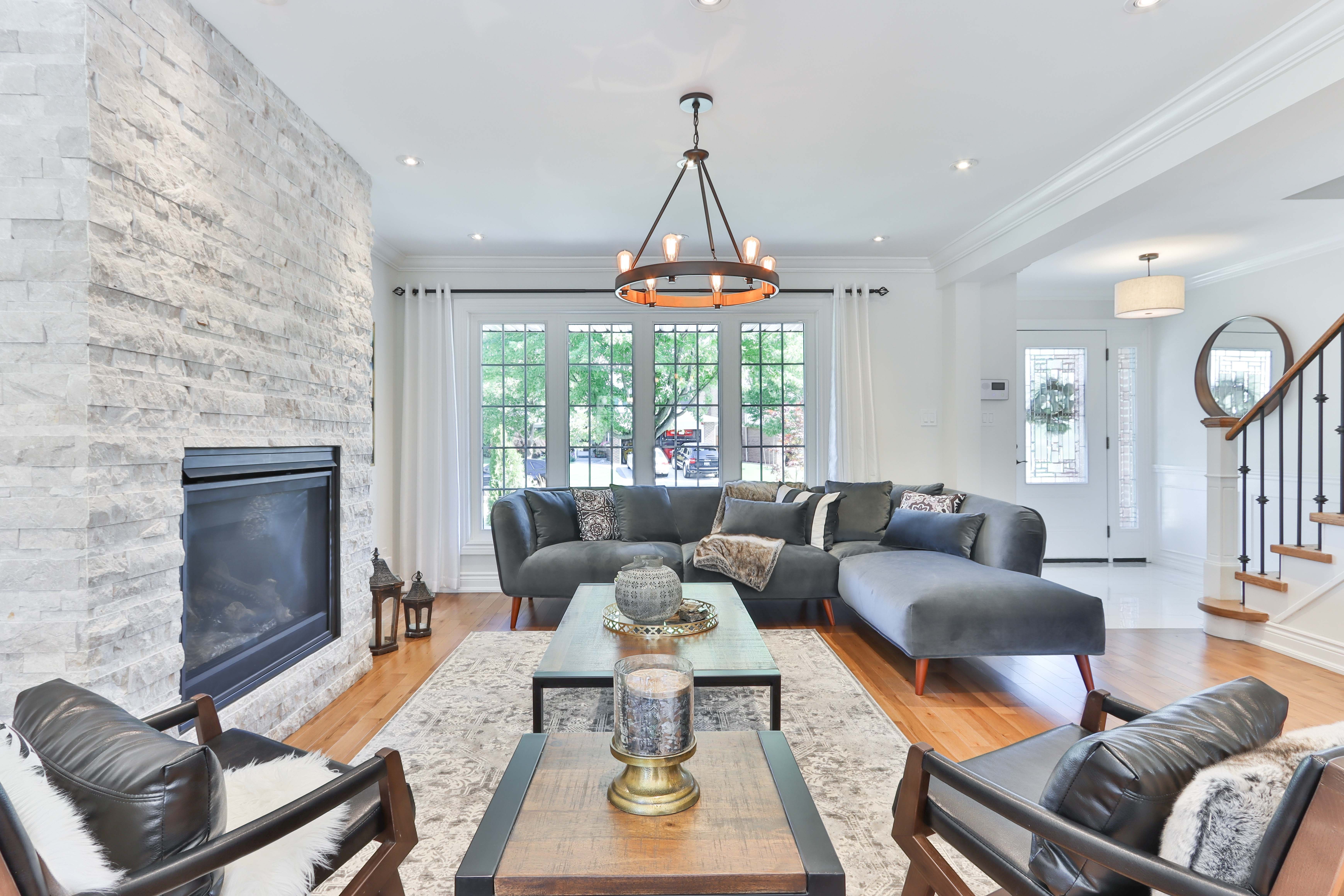 Staged living room with a stone fireplace wall, gray sectional, leather chairs, and large windows.
