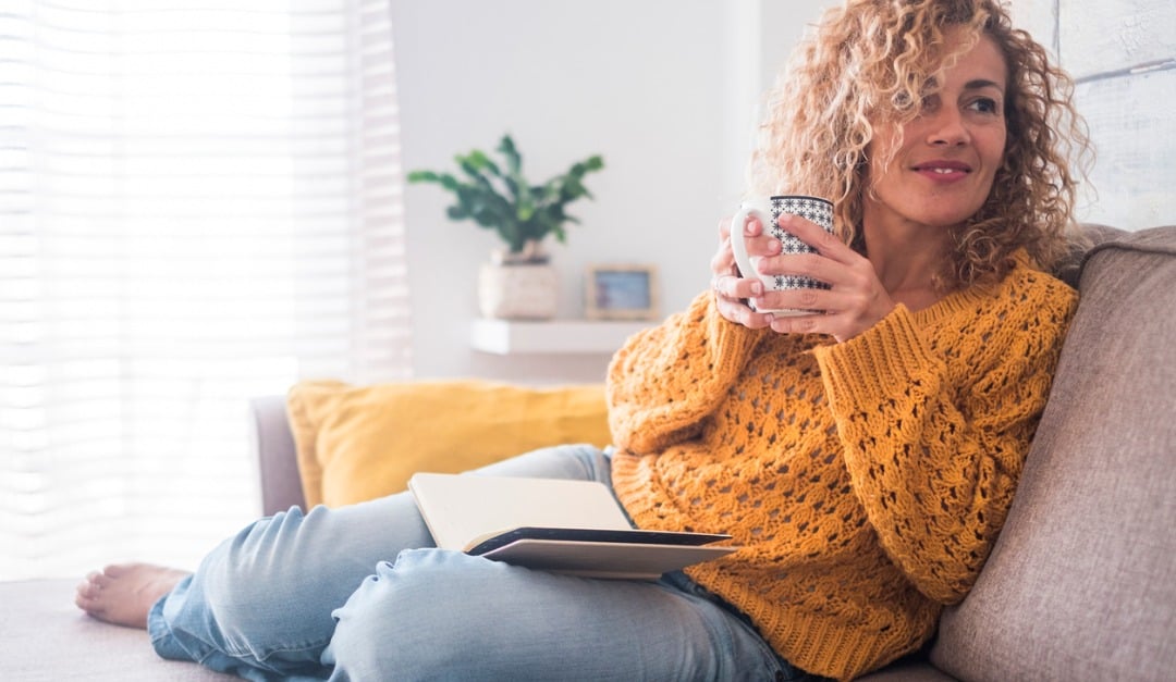 Woman relaxing on a sofa with a tablet on her lap and a mug in her hands.