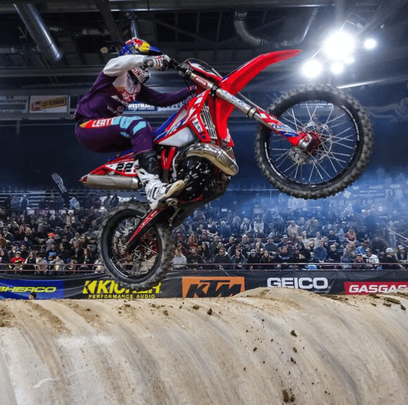 Motocross rider jumping a red dirt bike over a dirt ramp in a crowded indoor arena.