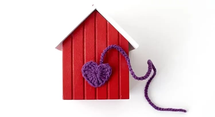 A small red house with a white roof and a purple heart-shaped decoration attached by a string. This image symbolizes warmth and love associated with home.