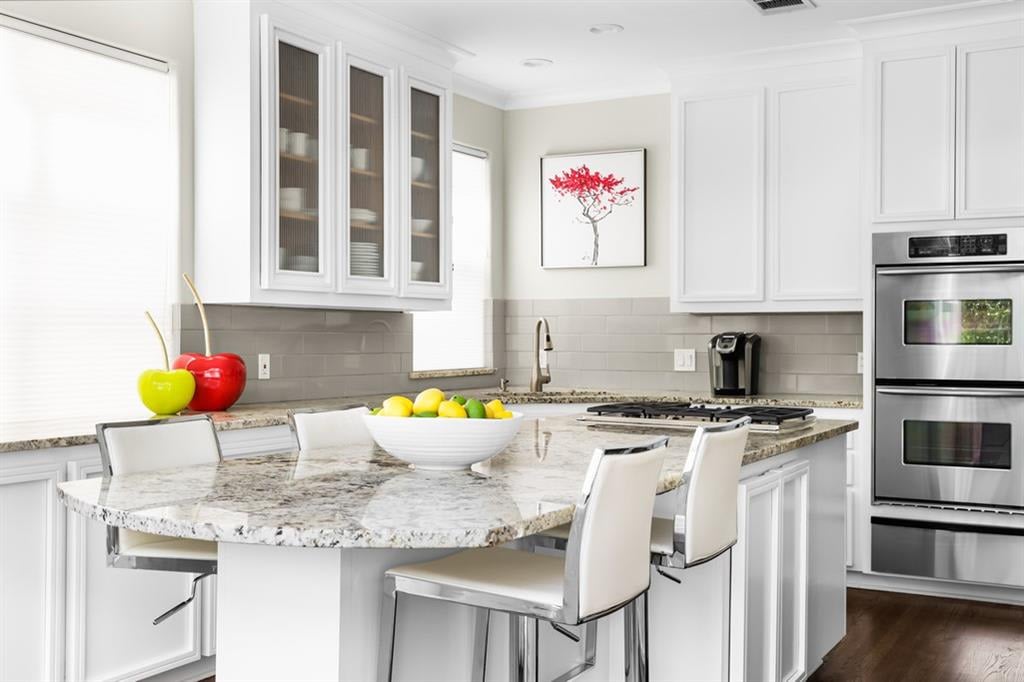 Bright white kitchen with a granite island, glass-front cabinets, and stainless-steel double oven.