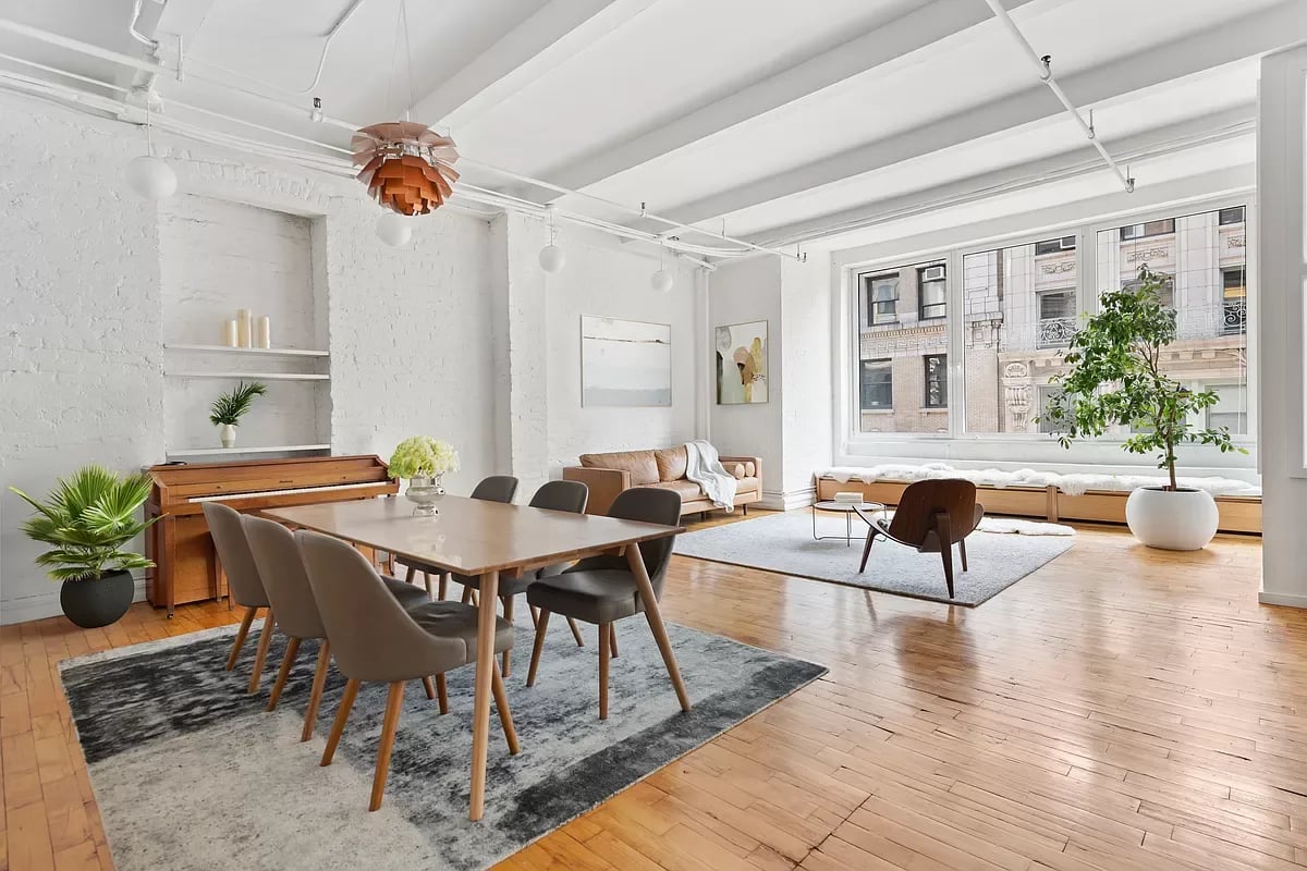 Open-plan living and dining room with white brick walls, wood floors, piano, and large windows.