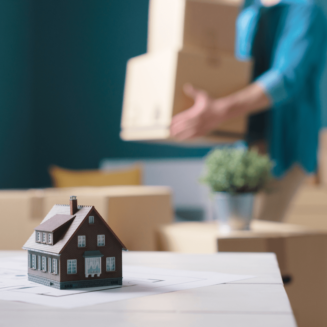 Miniature house on floor plans as a person carries a moving box in the background.