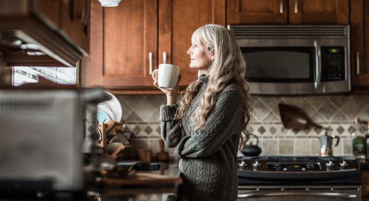  A woman drinking coffee in a kitchen with dark wooden cabinets and a stainless steel stove in the background. She appears to be enjoying her morning drink.