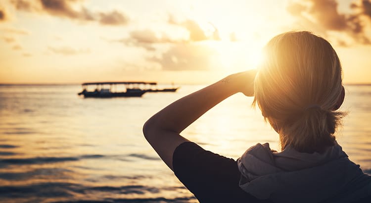 A person standing by the water, looking out towards the horizon at sunset. The sun is low in the sky, casting a golden glow over the scene, with a boat in the distance on the water.
