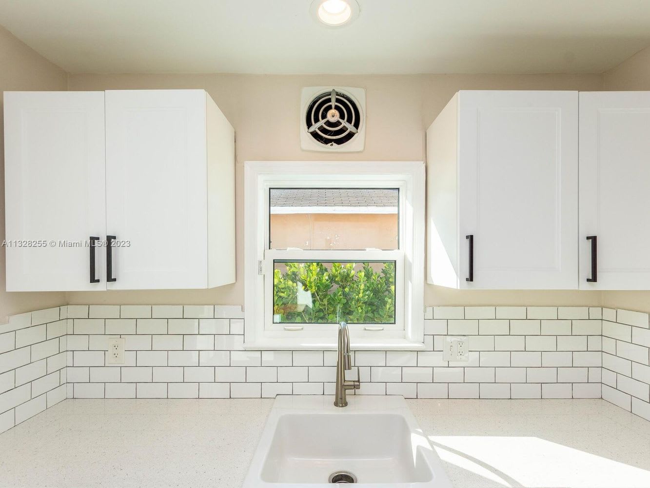 Kitchen with white cabinets and subway tile backsplash, centered sink beneath a window overlooking greenery.