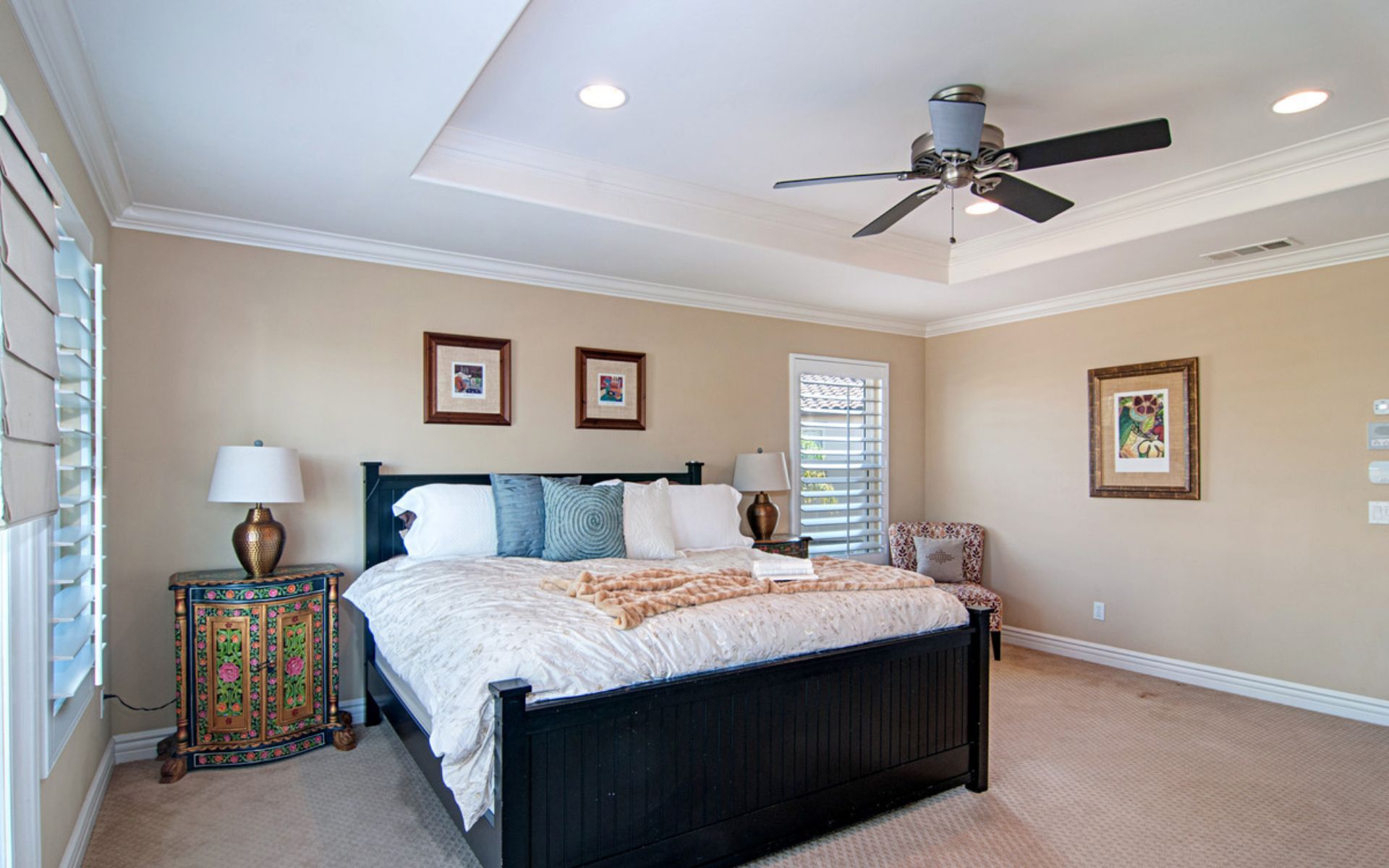 Bedroom with a dark wood bed, colorful bedside cabinet, shuttered windows, and ceiling fan.